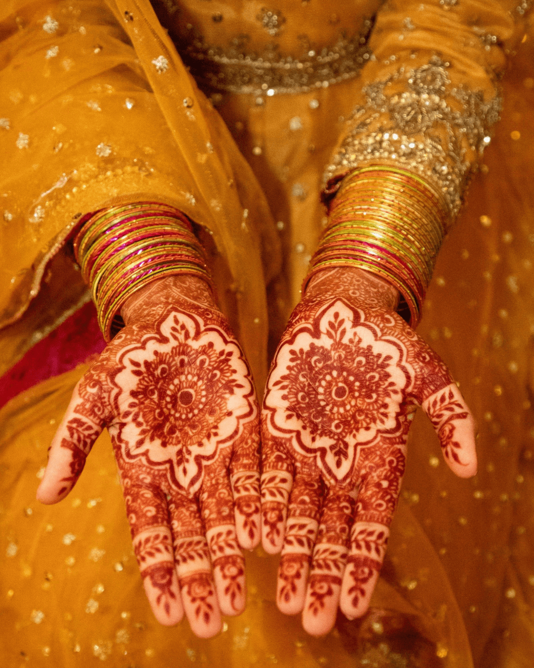 Henna-covered palms framed by gold and pink bangles against a mustard outfit