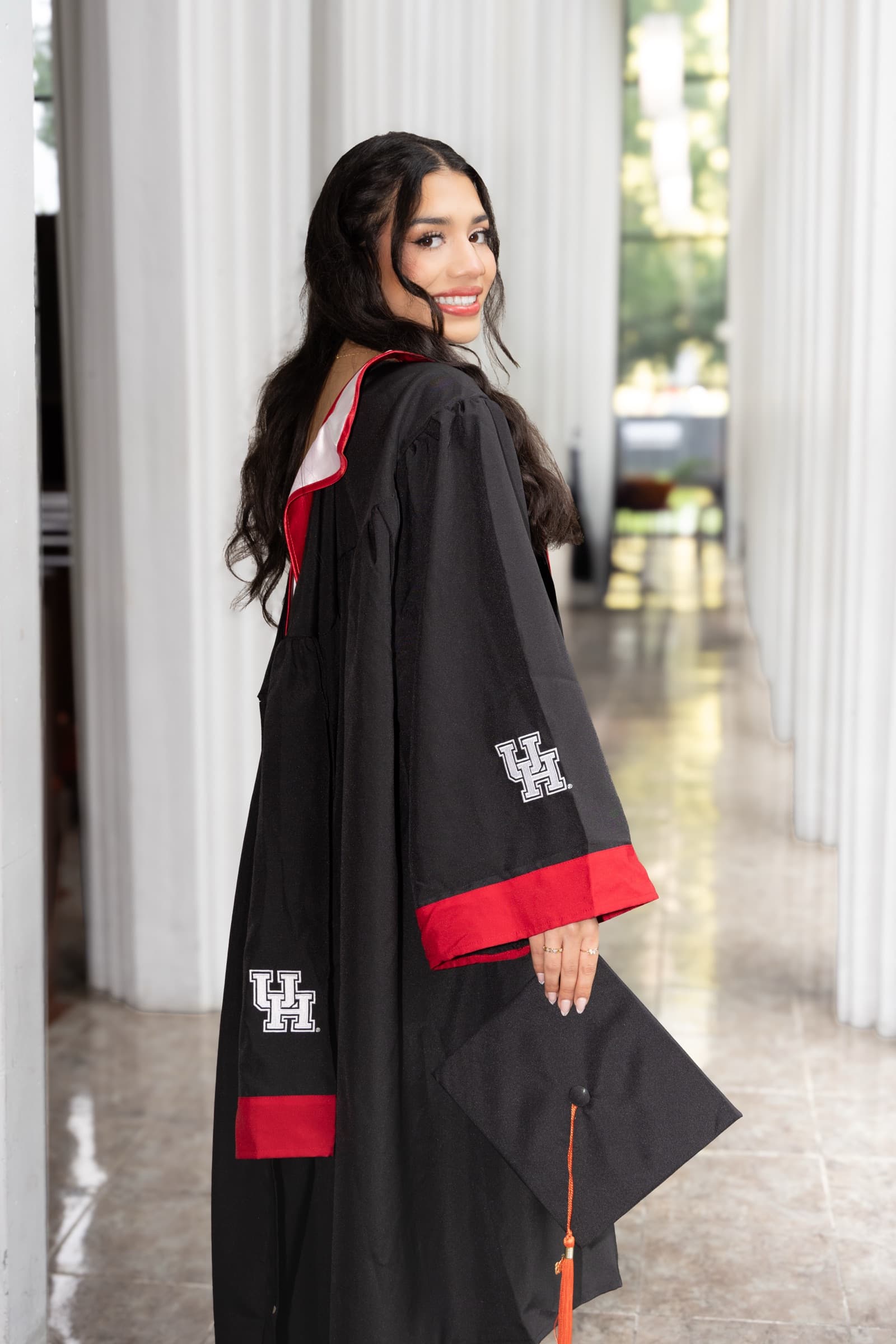 Graduate in cap and gown glancing back over her shoulder in a columned hallway