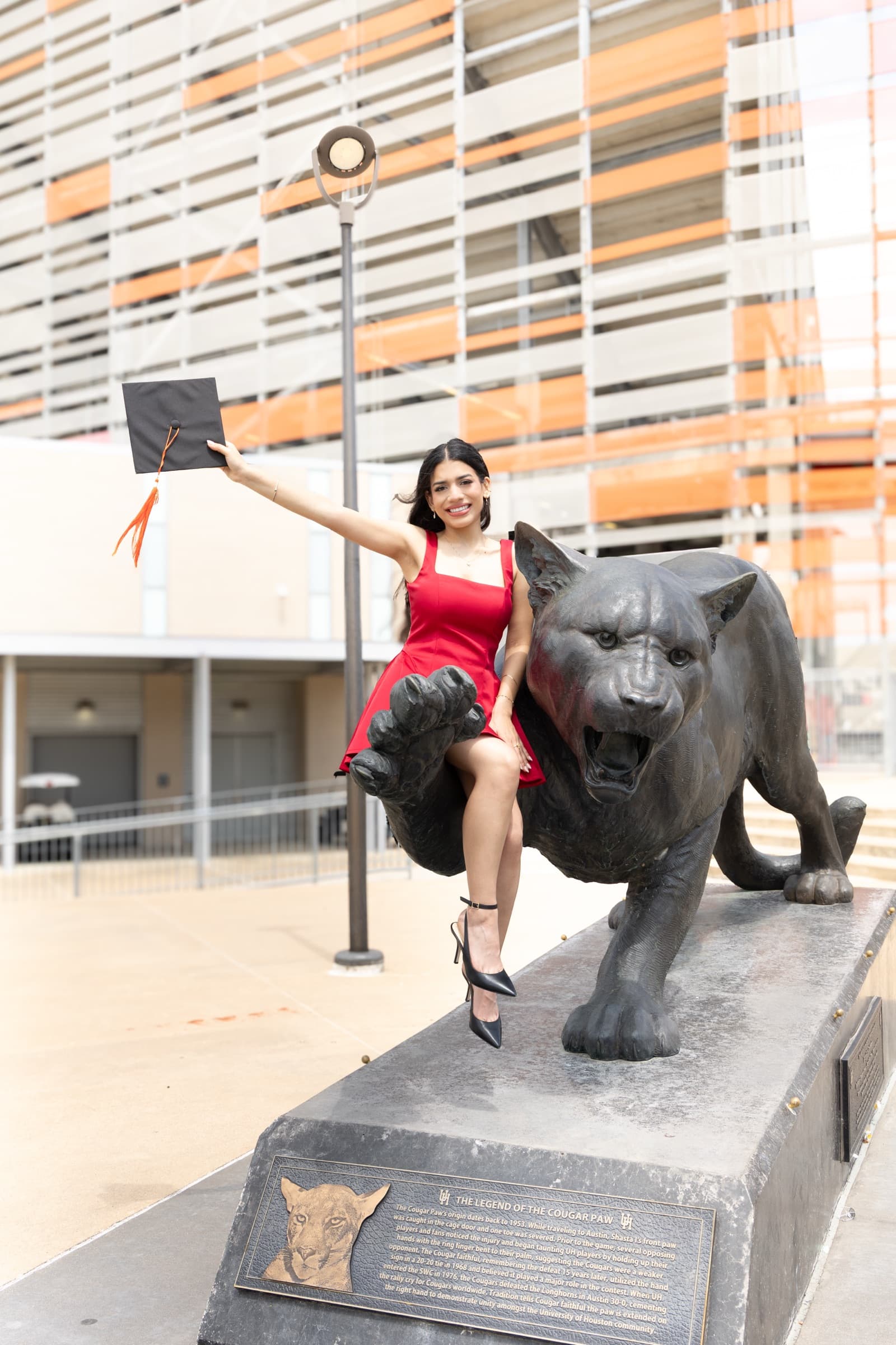 Graduate in a red dress sitting on the University of Houston Cougar statue, holding her cap