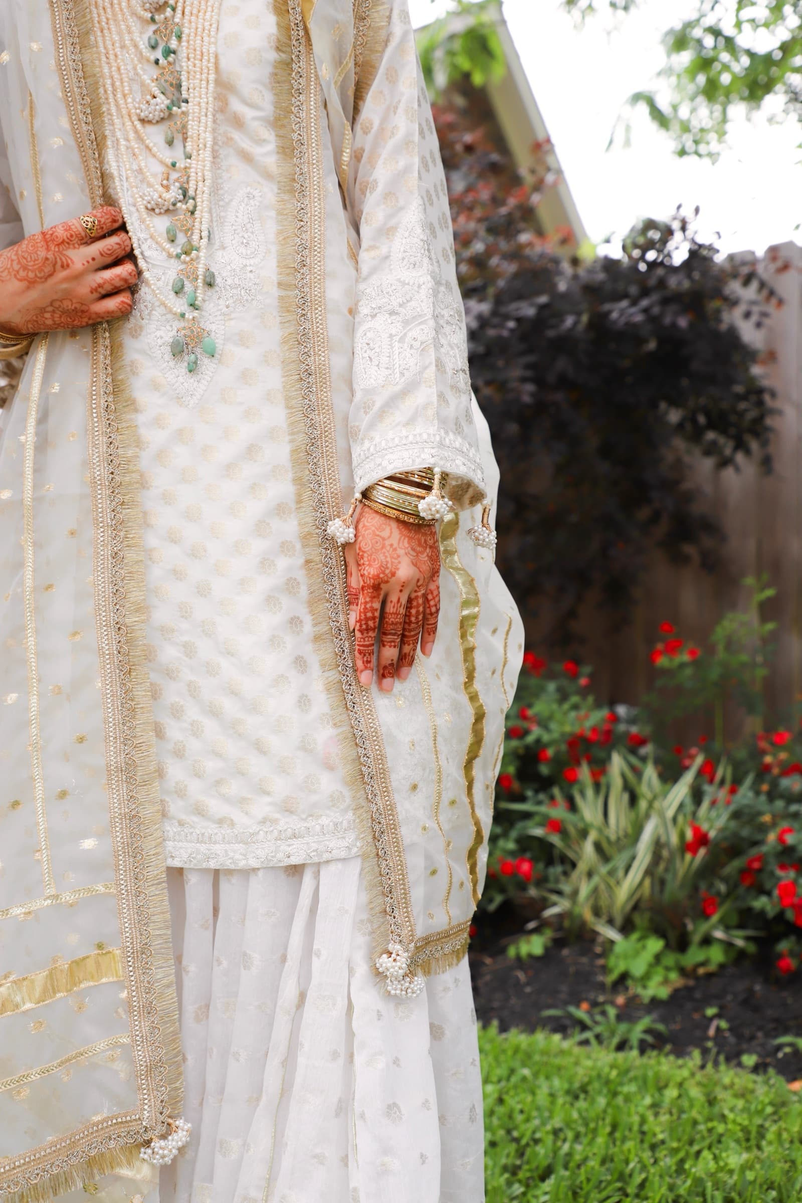 Bride in an ivory embroidered outfit with henna-adorned hands standing in a garden