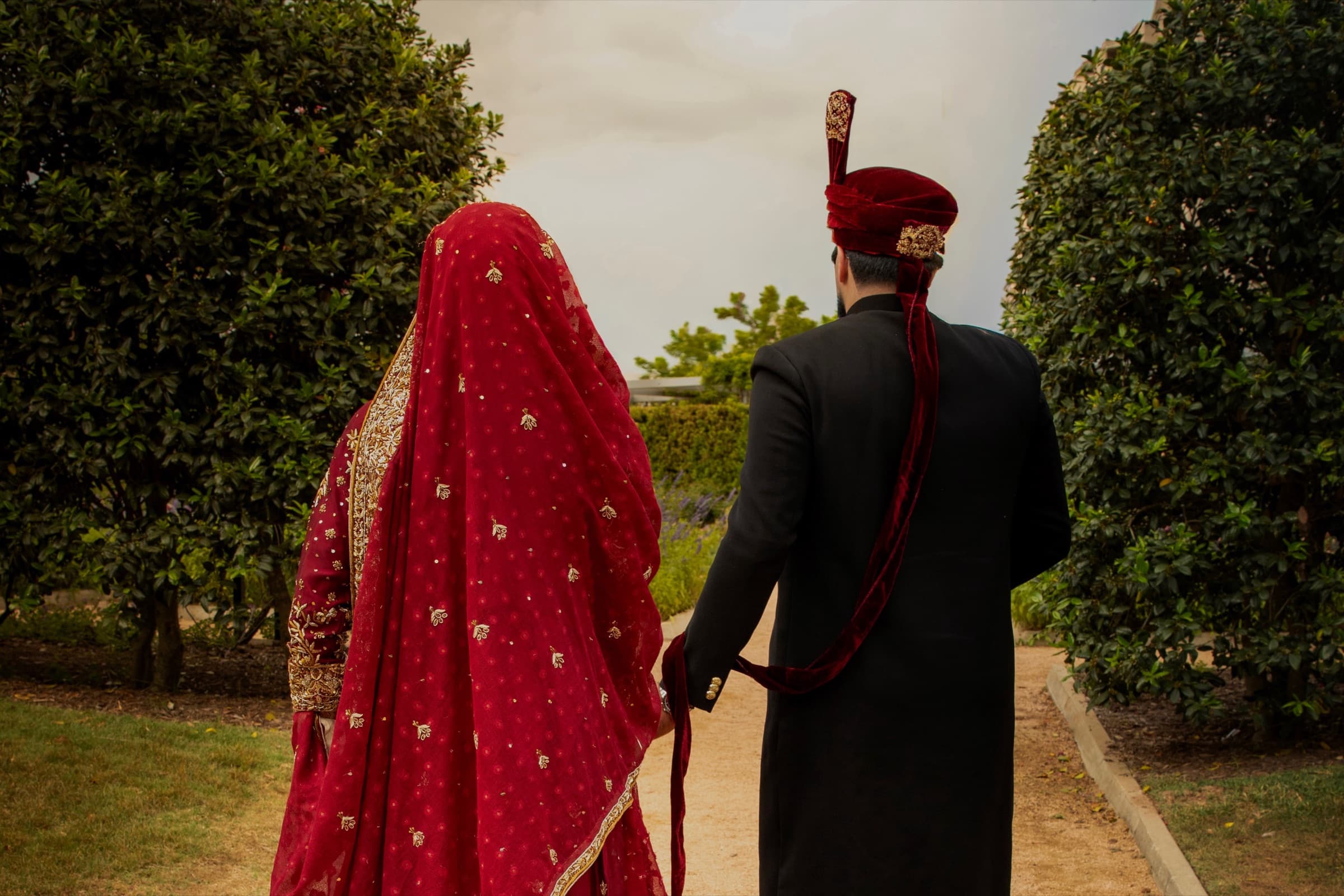 Bride in a red embroidered dupatta and groom in a black sherwani with maroon turban walking through a garden