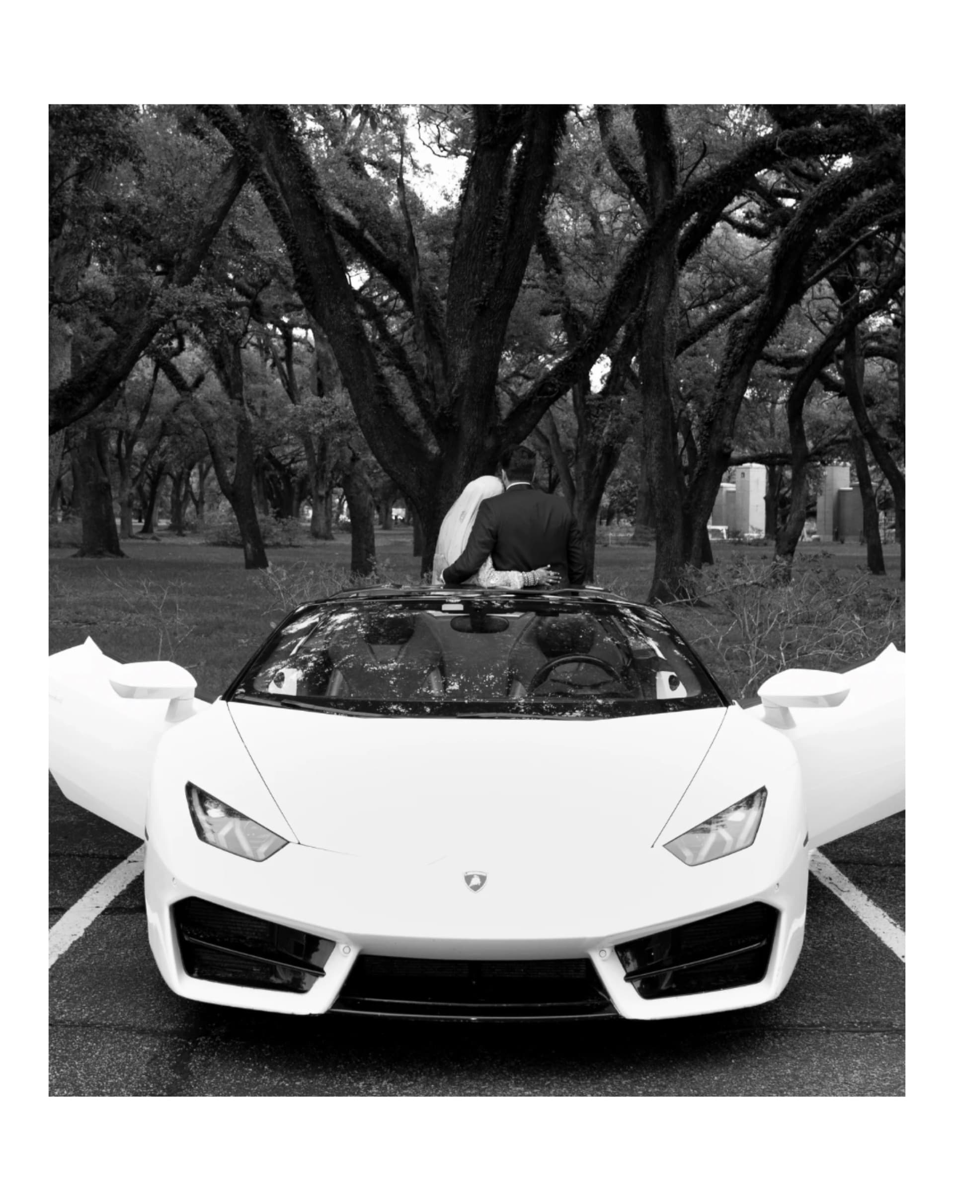 Black and white portrait of a bride and groom standing through the open doors of a sports car beneath oak trees